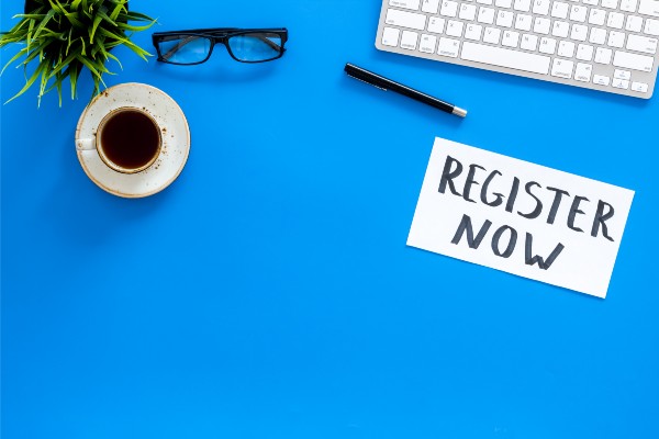 Register now sign on a blue background next to glasses, a coffee cup, a pen, and a keyboard.