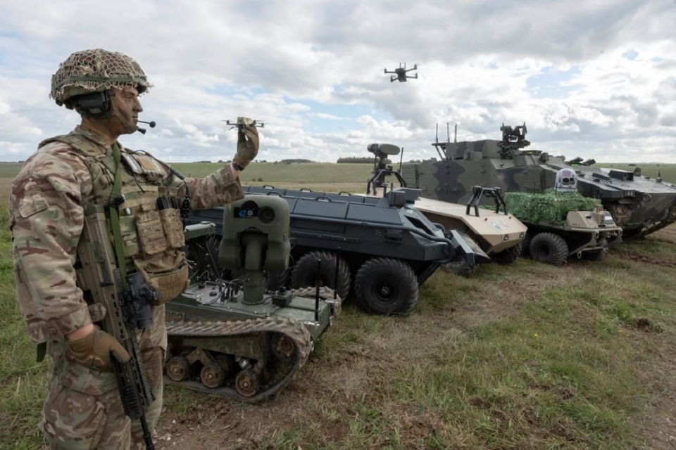A solider in British Army uniform at the Army Warfighting Experiment on Salisbury Plain holding a drone and standing next to different Army vehicles.