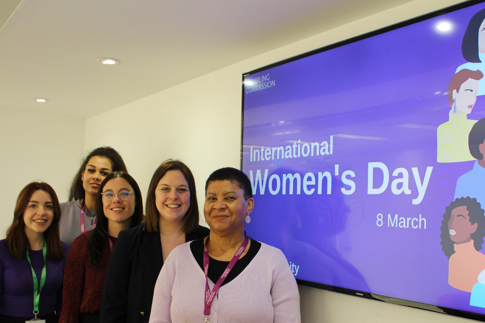 Gambling Commission comms team, consisting of five women, standing in front of a digital screen displaying graphics and text for International Women's Day - 8 March