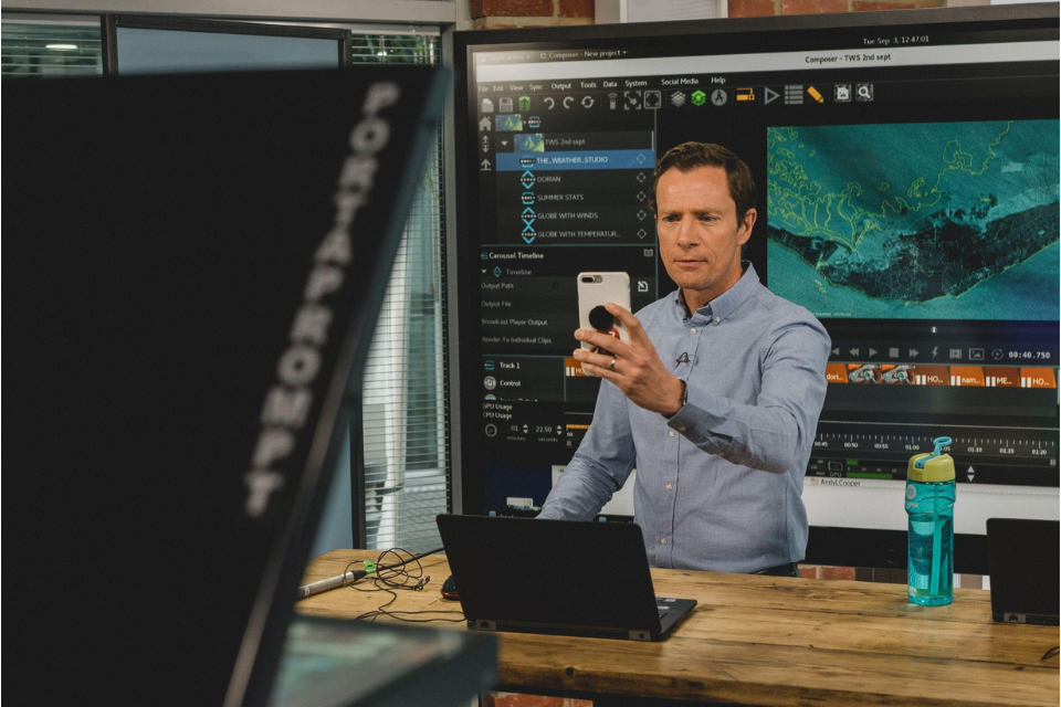 A person standing behind a wooden table upon which there is a laptop. The person is holding up their mobile phone in front of them. There is a large digital screen in the background