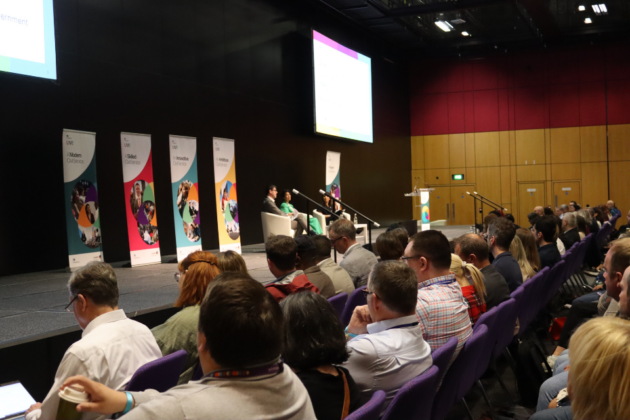 A stage with a seated panel of guest speakers presenting at Civil Service Live (CSL) in front of a large audience. There is two digital screens behind the panel and five CSL roll up banners with them on the stage.