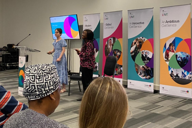 Jessica Ball (left) and Funke Oham (right) standing in front of a seated audience at Civil Service Live (CSL) 2024 in London. There is a lectern in front of them and four CSL roll up banners behind them.