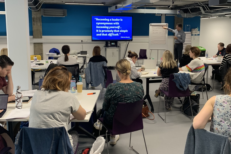 A room with people sitting at desks watching a presentation at the front of the room, where there is a blue digital screen with white writing and a man pointing to a flipchart