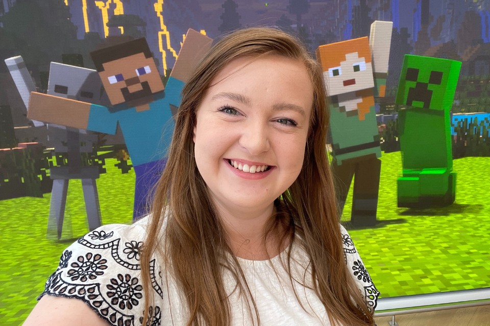 A selfie image of Abby Wade, GCS Applied Innovation Manager, at the Microsoft Experience Centre in London. Abby is smiling at the camera and there is a scene of Minecraft characters in the background.