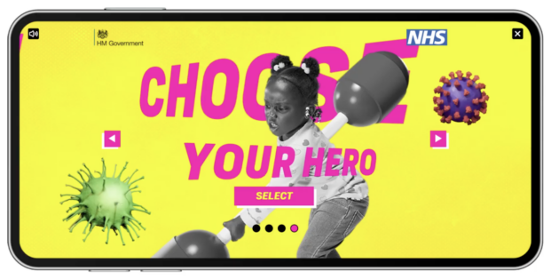 A vibrant NHS and HM Government poster designed in the style of a mobile video game character selection screen. The background is bright yellow with large magenta text reading “CHOOSE YOUR HERO” and a “SELECT” button below. At the centre is a black and white photo of a young girl holding a toy pugil stick, as if preparing for battle. Illustrations resembling flu particles appear on either side of her. There are navigation arrows and a row of dots indicating multiple selection screens.